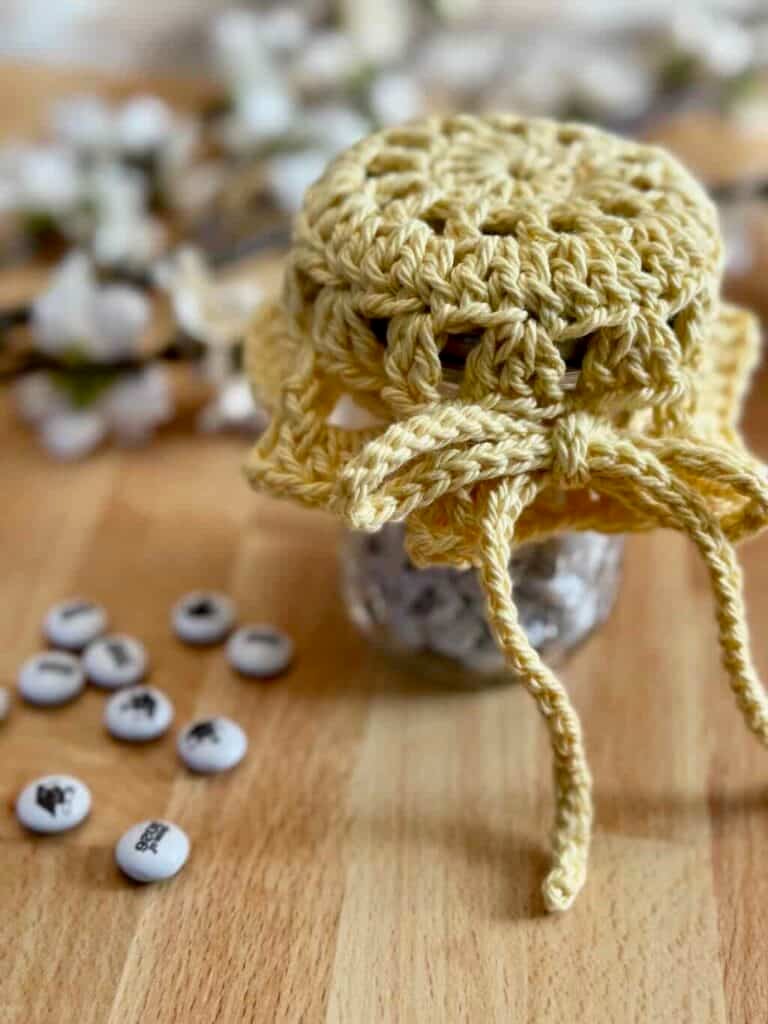 A mason jar crochet cover doily on top of a mason jar filled with candy, on a wooden background with flowers
