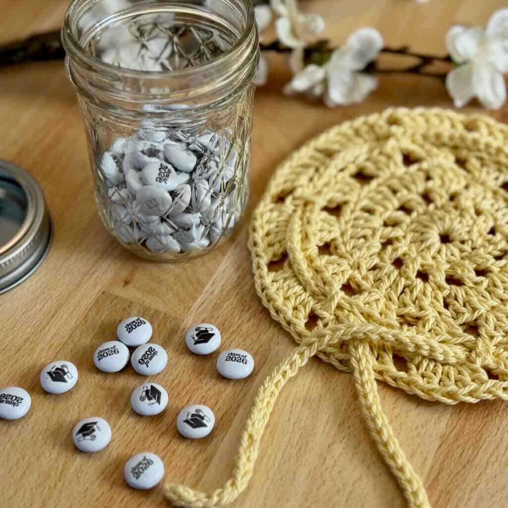 A photo of a mason jar filled with candy alongside a mason jar crochet cover doily on a wooden background with flowers