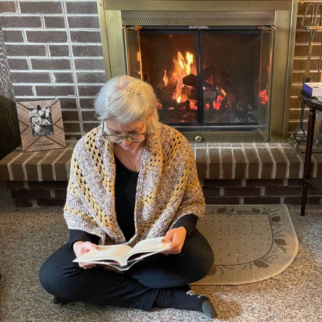 Photo of the granny stitch shawl wrapped around the shoulders of a woman reading by the fireplace